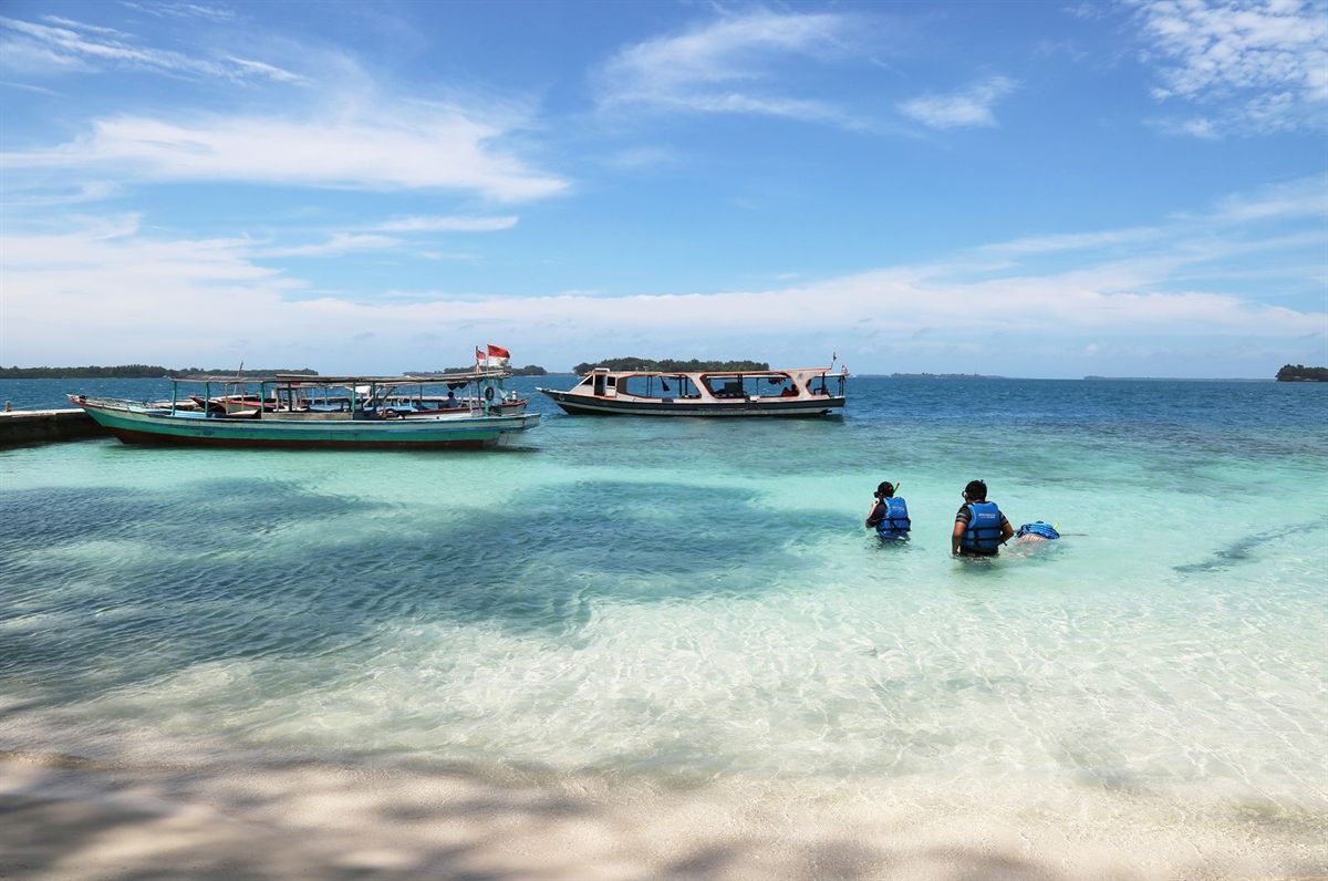 Small boats moored off a white sand beach with people wading in shallow water